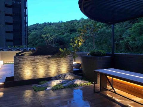 蕭東駿 - 夜幕下的景觀以水瀑牆為視覺焦點，柔和燈光從牆面與步道間流動，勾勒出溫暖而靜謐的氛圍。搭配岩石造景、植栽層次與木質座椅的光影細節，整體空間展現沉穩且富質感的夜間庭園韻味，營造出放鬆、舒適的戶外休憩場景。