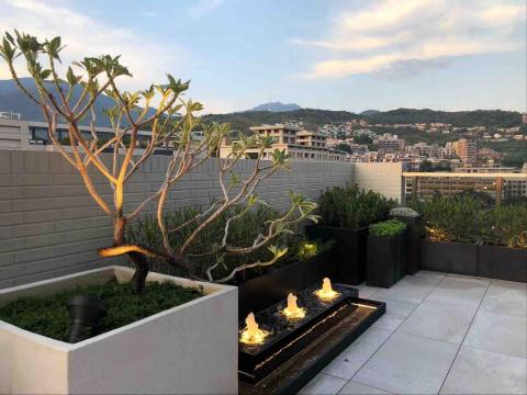 蕭東駿 - 這處約20坪的露臺以架高地磚鋪設，呈現簡潔大方的戶外空間。中央設置的噴水池與周圍植栽相互呼應，營造流動水景與綠意並存的放鬆氛圍；搭配木質座椅與盆栽配置，使整體景觀更具層次與生活感，成為城市中難得的靜謐角落。