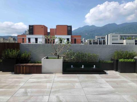 蕭東駿 - 這處約20坪的露臺以架高地磚鋪設，呈現簡潔大方的戶外空間。中央設置的噴水池與周圍植栽相互呼應，營造流動水景與綠意並存的放鬆氛圍；搭配木質座椅與盆栽配置，使整體景觀更具層次與生活感，成為城市中難得的靜謐角落。