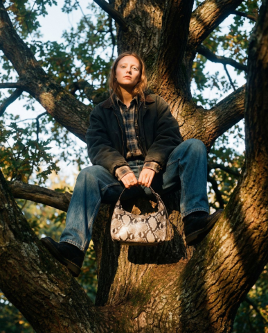 FpVisualAi - 雙手輕握著一款蛇紋圖案的手提包，包身採用銀灰與米白交織的亮面蛇皮紋理，在陽光下反射出細膩的光澤，與她樸實的穿搭形成有趣的對比。
她整個人倚坐在粗壯的樹枝間，雙腿自然分開，一腳踩在下方樹幹上，陽光從側面灑落，在樹皮和她身上留下斑駁而溫暖的光影。背景是茂密的樹葉與藍天，整體畫面充滿秋日午後的寧靜與自由氣息，色調以大地色、木紋棕和藍綠為主，氛圍自然且極具時尚感。